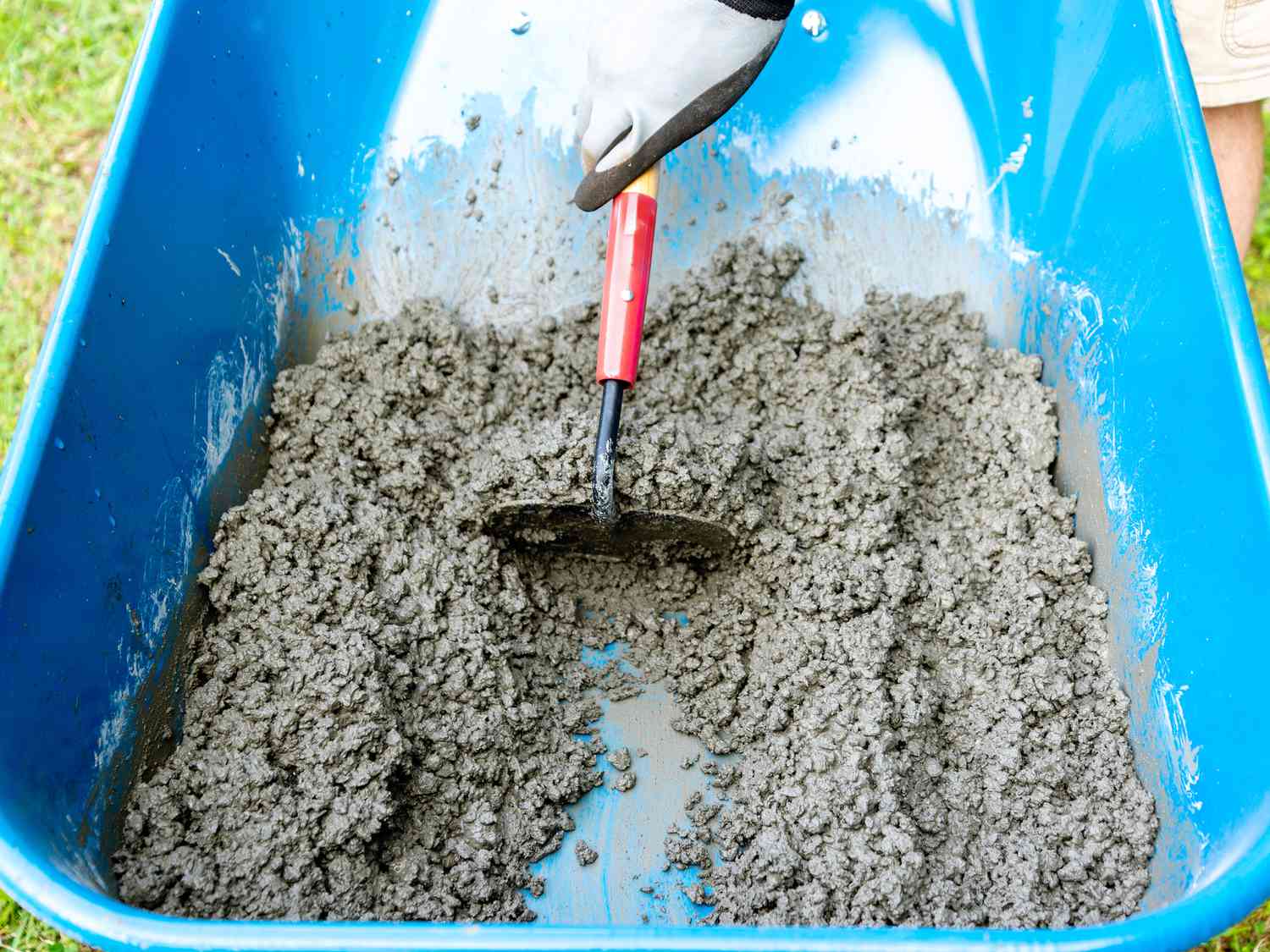 Une personne mélange du béton avec une houe à béton dans une brouette bleue.