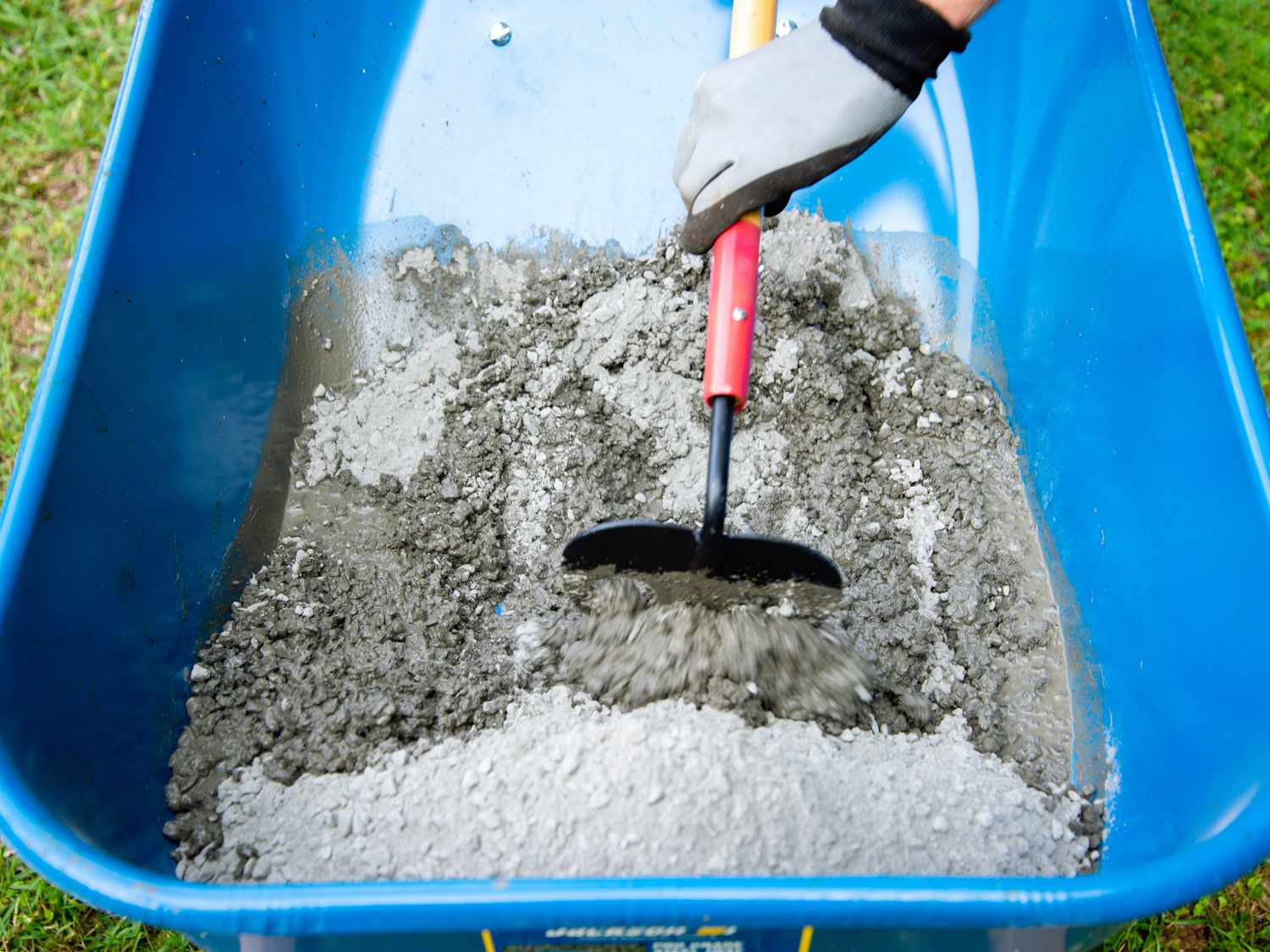 Une personne utilise une houe à béton pour mélanger le béton dans une brouette bleue.