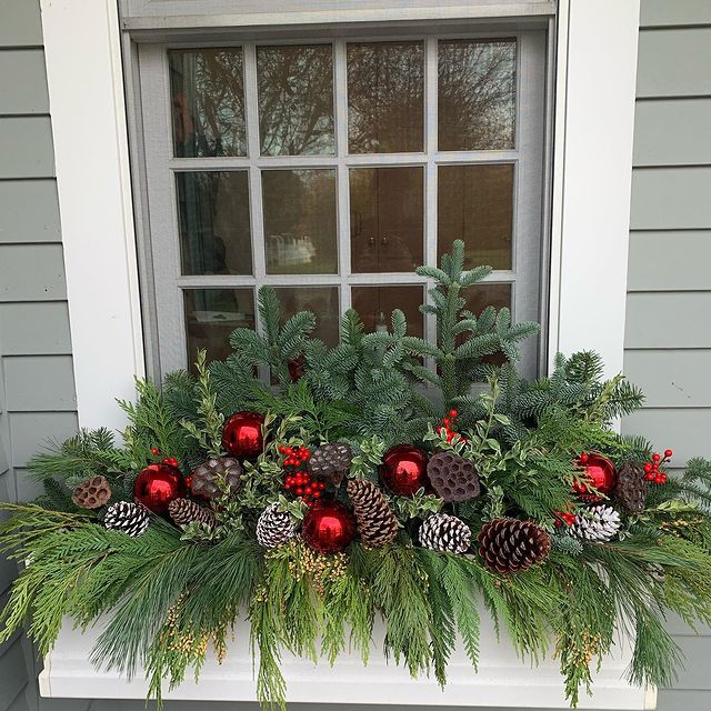 jardinière blanche sur maison grise avec branches de conifères, pommes de pin, baies rouges et ornements en verre rouge